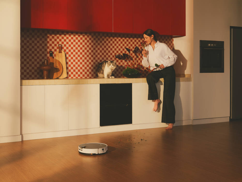 Kobieta siedzi na blacie kuchennym obok kota, trzymając w ręku warzywa. Na podłodze widać biały robot sprzątający oraz rozsypane liście.