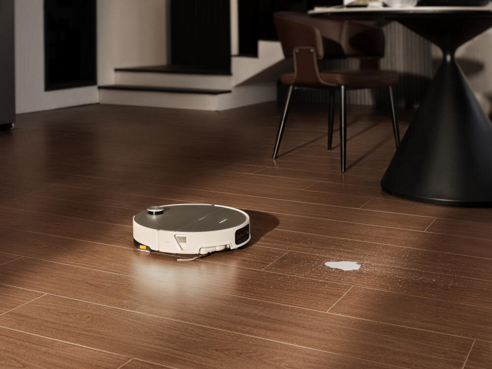 Na drewnianej podłodze znajduje się biały robot sprzątający. W tle widoczny jest stół z krzesłem.