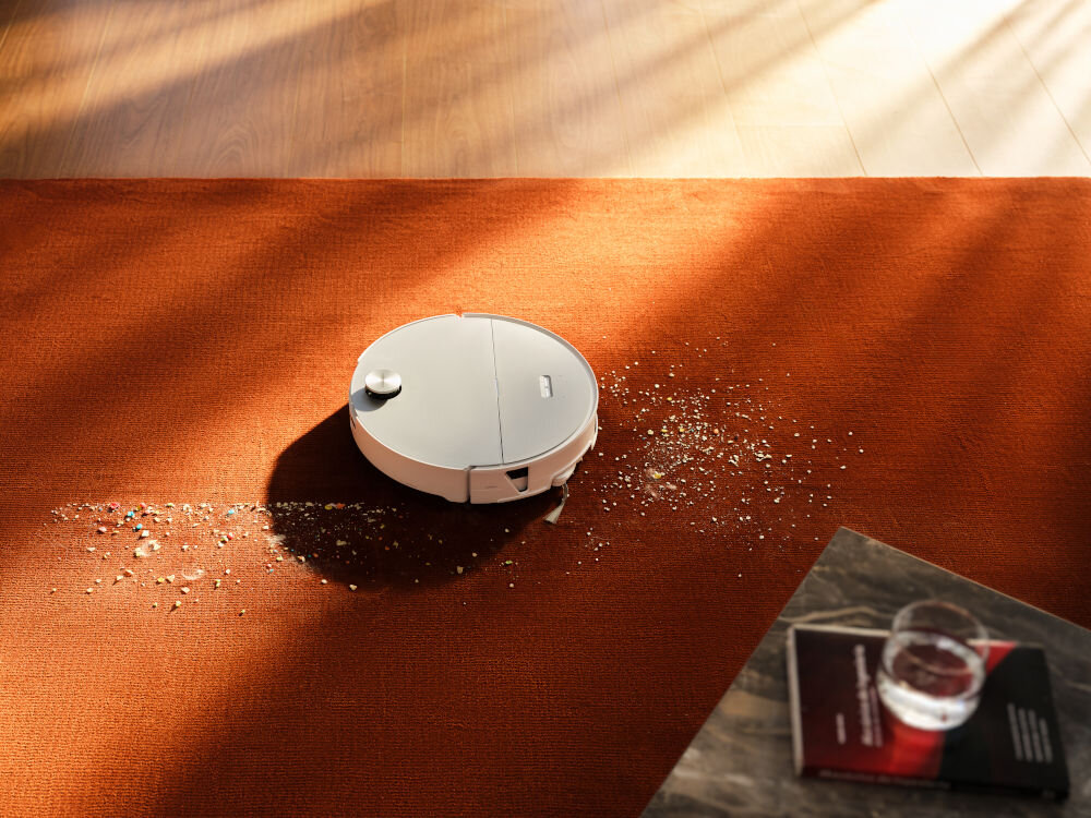 Na pomarańczowym dywanie znajduje się biały robot sprzątający, który odkurza rozsypane okruchy. Obok leży książka i szklanka z wodą na stoliku.