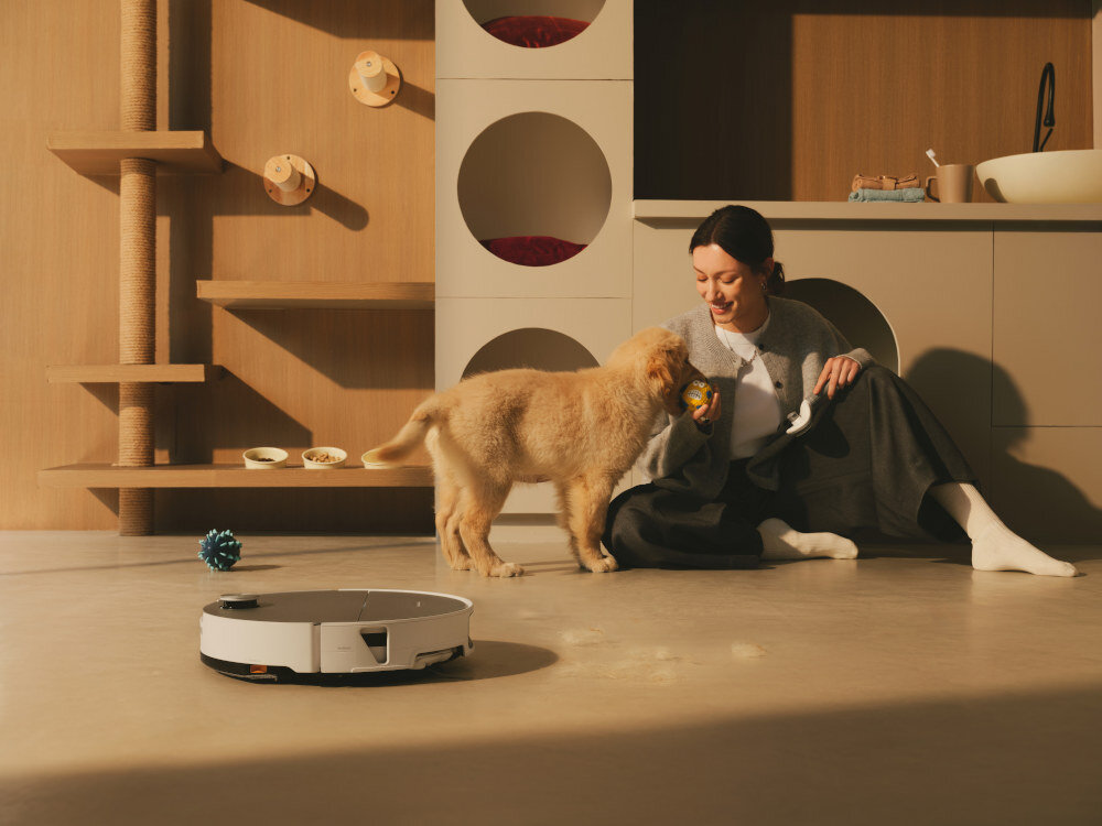 Na podłodze znajduje się biały robot sprzątający. Obok siedzi osoba bawiąca się z psem w jasnym pomieszczeniu z meblami i drapakiem dla kota.