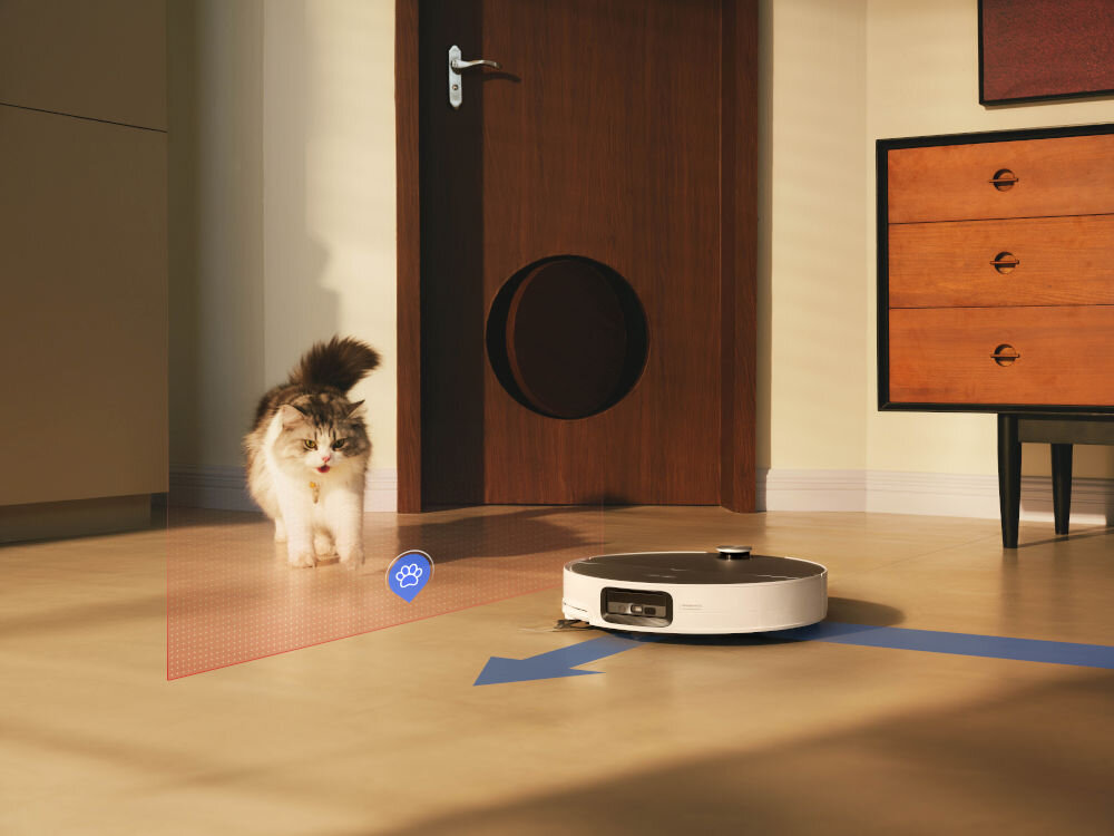 Na podłodze znajduje się biały robot sprzątający, a obok niego idzie kot. Na podłodze widoczna jest niebieska strzałka oraz symbol łapy w niebieskim kółku.
