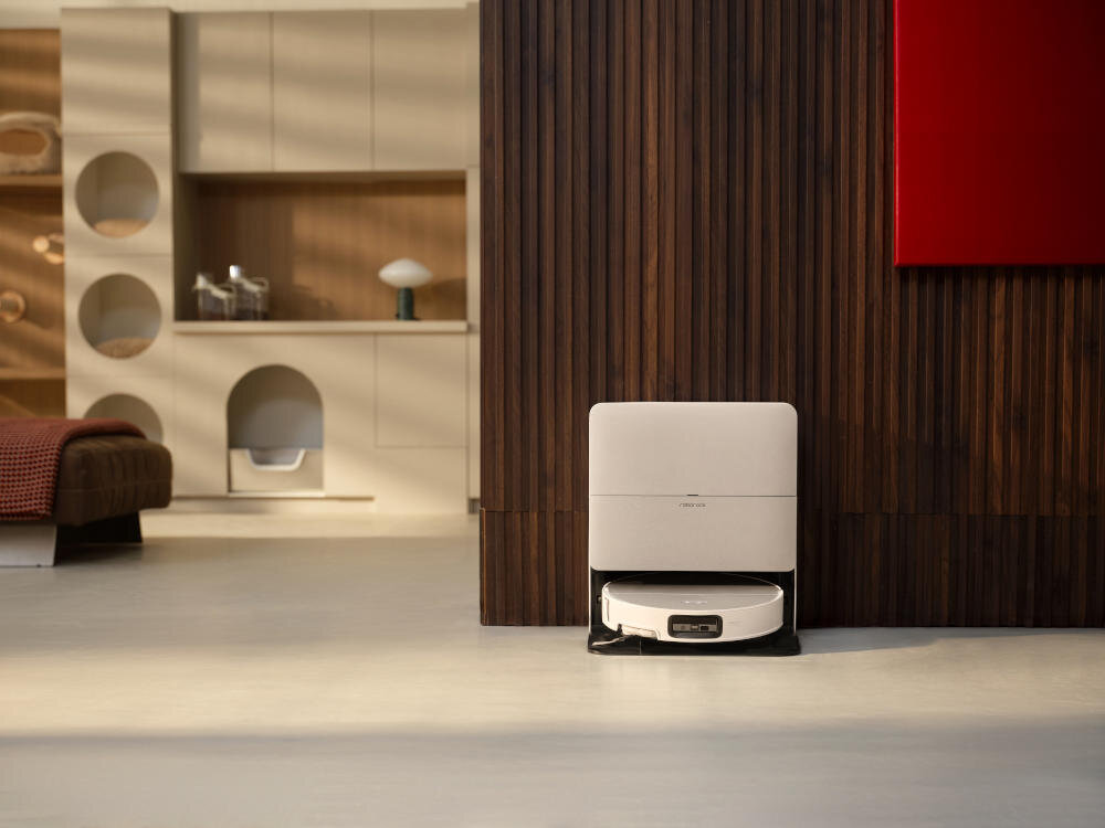 Na obrazie widoczny jest biały robot sprzątający umieszczony w stacji dokującej na tle drewnianej ściany. W tle znajduje się nowoczesne wnętrze z półkami i czerwonym obrazem na ścianie.