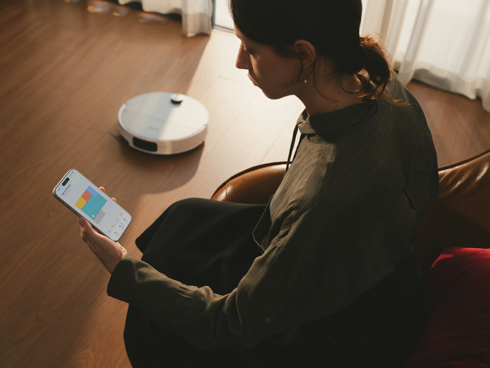 Osoba siedzi na kanapie, trzymając w ręku smartfon z aplikacją pokazującą kolorowe wykresy. Na podłodze widać biały robot sprzątający.