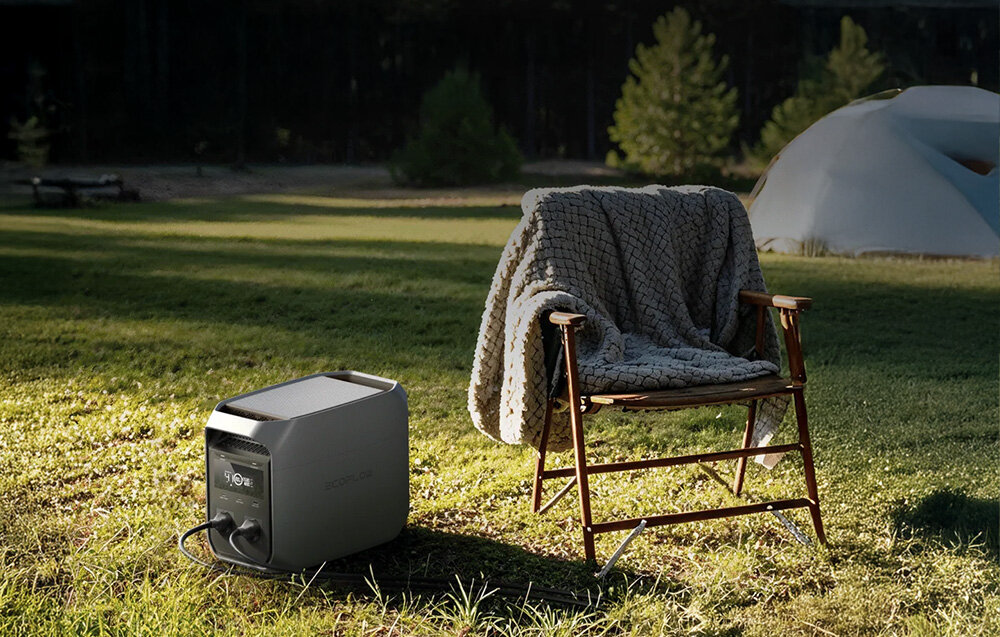 Na trawie stoi stacja zasilania obok krzesła z narzuconym kocem, w tle widoczny jest namiot i drzewa.
