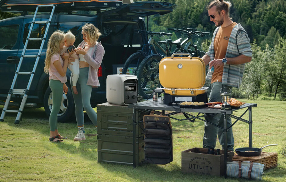 Na trawie stoi rozłożony stół z grillem i stacją zasilania ECOFLOW Delta 3 Classic 1000 Wh. W tle widać samochód z rowerami i drabiną, a obok stoi grupa osób.