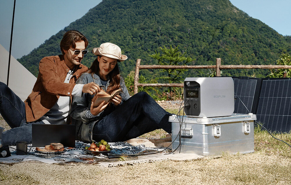 Dwie osoby siedzą na kocu na trawie, obok nich znajduje się stacja zasilania ECOFLOW Delta 3 Classic 1000 Wh umieszczona na metalowej skrzyni. W tle widoczne są panele słoneczne i zielone wzgórza.