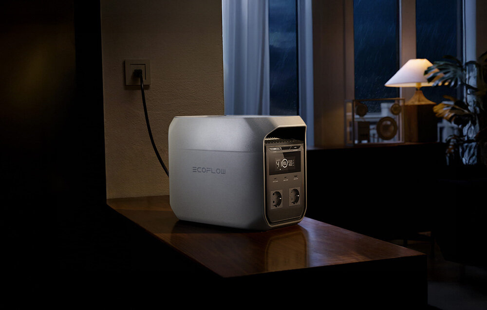Na drewnianym stole stoi stacja zasilania ECOFLOW z wyświetlaczem i gniazdami. W tle widać okno z zasłonami i lampę stołową.