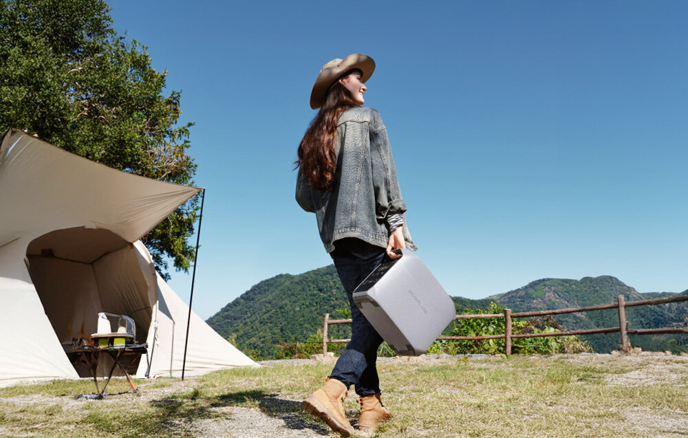 Osoba w kapeluszu trzyma przenośną stację zasilania na tle namiotu i górskiego krajobrazu. Na stacji widoczny jest napis \'ECOFLOW\'.
