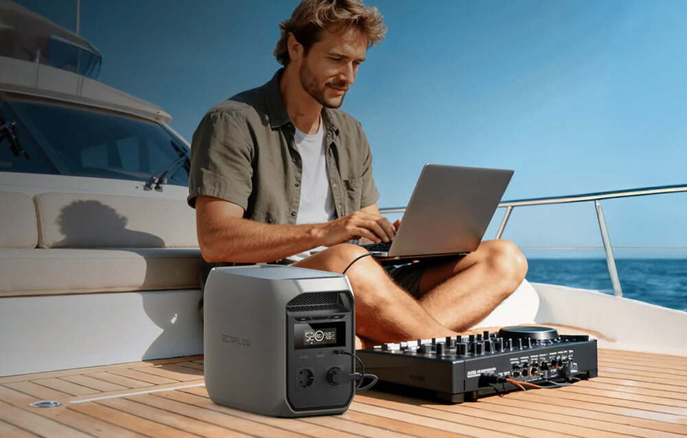 Osoba siedzi na pokładzie łodzi, używając laptopa, obok znajduje się stacja zasilania ECOFLOW Delta. W tle widać fragment jachtu i niebo.