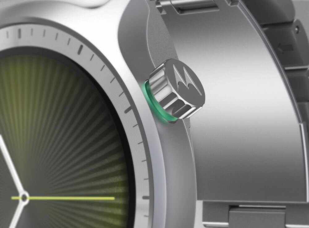 Zbliżenie na metalową kopertę zegarka z widocznym przyciskiem z logo Motorola. Tarcza zegarka ma ciemnozielony gradient i cienkie wskazówki.