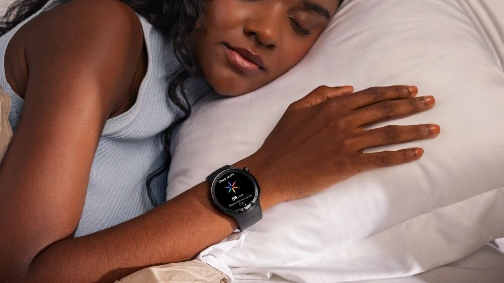 Osoba leży na poduszce, mając na nadgarstku czarny smartwatch z okrągłym wyświetlaczem. Na ekranie zegarka widoczny jest kolorowy wskaźnik i tekst.