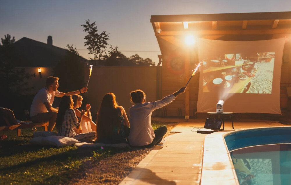 Projektor KIANO Vision 2 Smart TV Netflix YouTube screen mirroring telefon, projektor stojący na stoliku w plenerze, wyświetlający obraz na białym płótnie