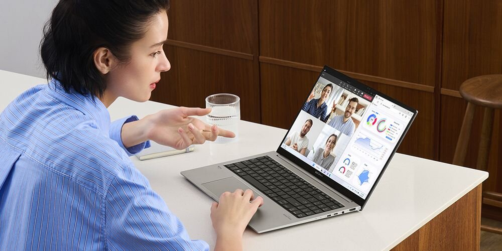 Kobieta w niebieskiej koszuli w paski używająca laptopa ASUS ZenBook podczas wideokonferencji, trzymająca szklankę wody i patrząca na ekran z uczestnikami spotkania online - funkcje poprawy jakości wideorozmów 