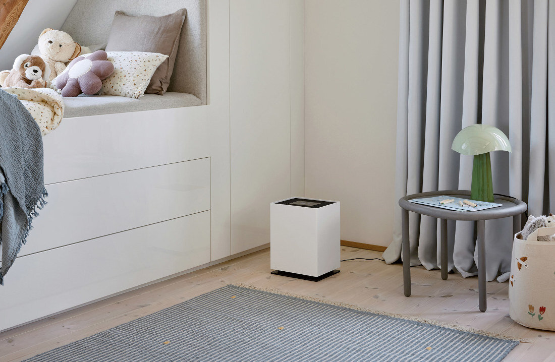 W jasnym pokoju dziecięcym znajduje się biały nawilżacz ewaporacyjny o prostokątnym kształcie. Obok stoi okrągły stolik z zieloną lampką, a na łóżku leżą pluszowe zabawki.