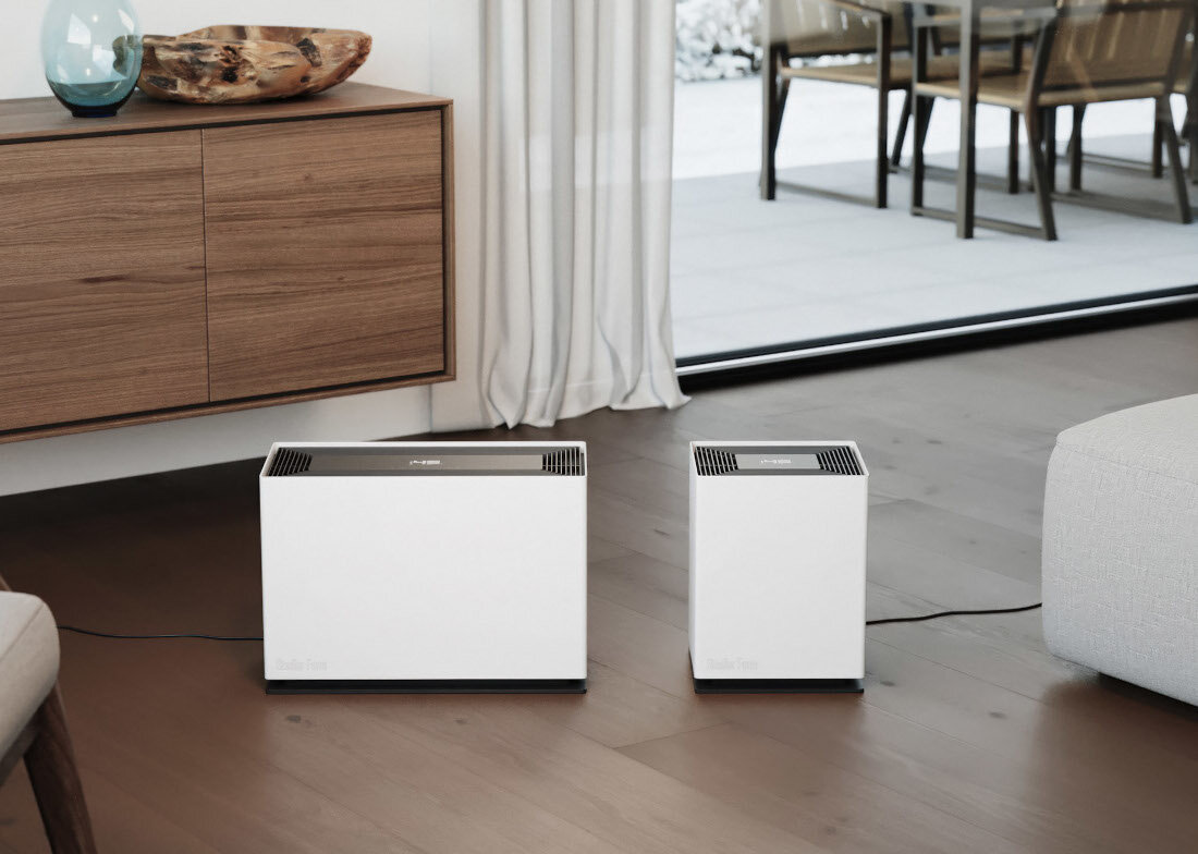 Na zdjęciu znajdują się dwa białe nawilżacze ewaporacyjne ustawione na drewnianej podłodze w nowoczesnym wnętrzu. W tle widoczna jest drewniana komoda, zasłony i przeszklone drzwi prowadzące na taras.