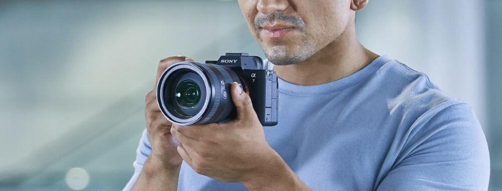 Osoba trzyma aparat bezlusterkowy marki Sony z widocznym obiektywem. Aparat jest czarny z logo Sony i symbolem α7 na korpusie.