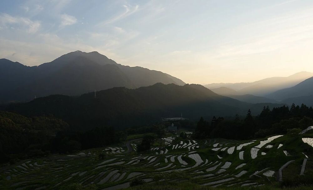 Krajobraz górski o zachodzie słońca z widocznymi tarasami ryżowymi na pierwszym planie. Niebo jest jasne z delikatnymi chmurami, a góry są ciemne na tle nieba.