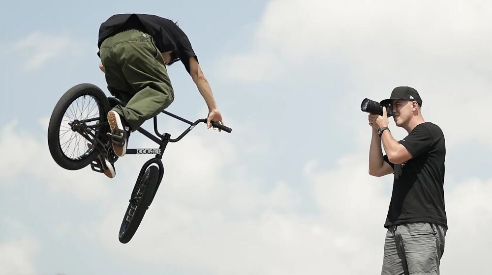Osoba na rowerze BMX wykonuje skok w powietrzu, podczas gdy inna osoba fotografuje tę scenę aparatem. W tle widoczne jest niebo z chmurami.