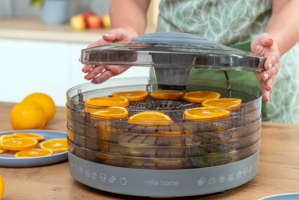 Osoba obsługuje suszarkę do żywności marki milla home, w której znajdują się plastry pomarańczy. Na urządzeniu widoczne jest logo \'milla home\'.