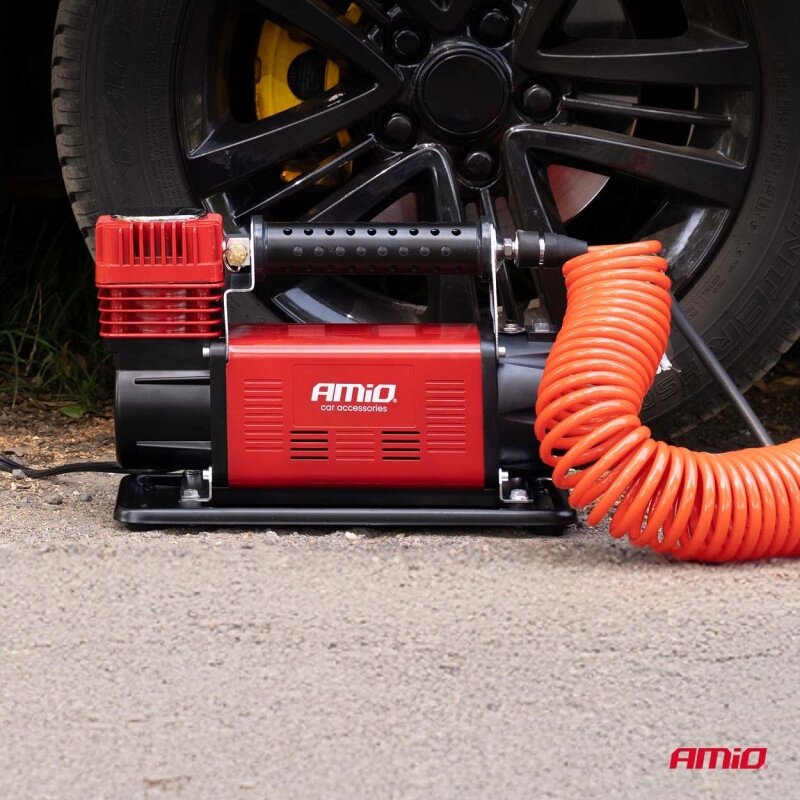 Na obrazie widoczny jest czerwony kompresor samochodowy z napisem \'AMiO car accessories\', obok którego znajduje się pomarańczowy spiralny wąż. W tle widać fragment czarnego koła samochodowego.