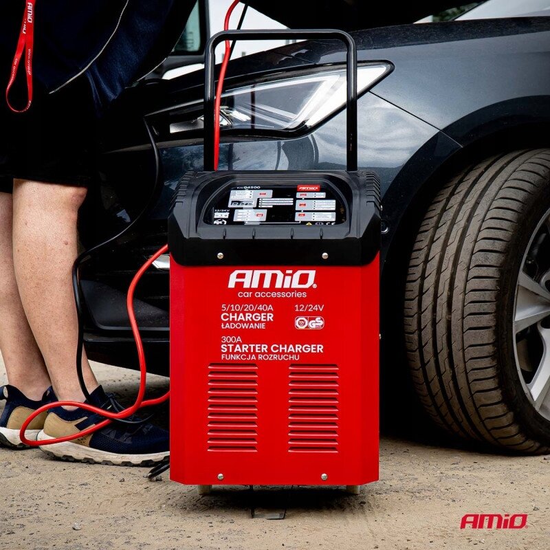 Na zdjęciu widoczny jest czerwony prostownik z rozruchem marki AMiO, podłączony do samochodu. Na urządzeniu znajduje się tekst: \'AMiO car accessories 5/10/20/40A CHARGER ŁADOWANIE 12/24V 300A STARTER CHARGER FUNKCJA ROZRUCHU\'.