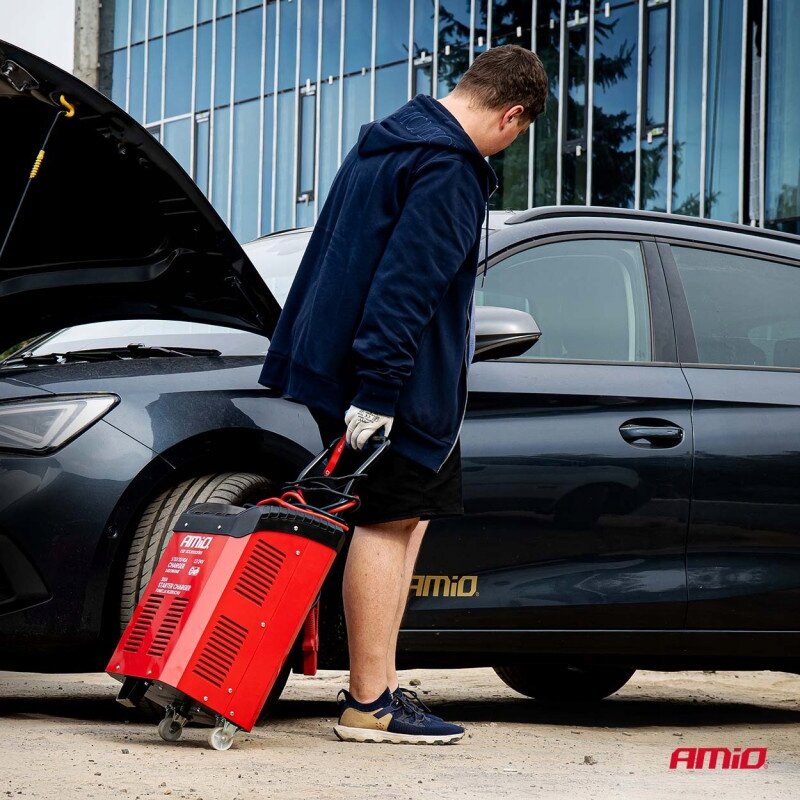 Osoba trzyma czerwoną ładowarkę samochodową AMiO obok otwartego samochodu. Na ładowarce widoczny jest tekst: \'AMiO BATTERY CHARGER\'.