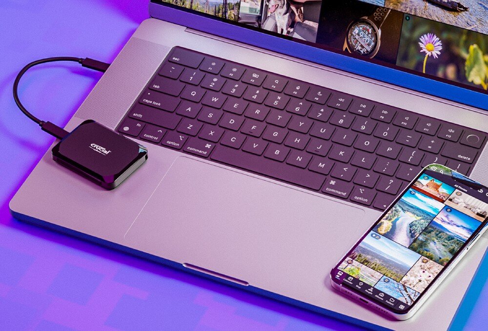 Dysk CRUCIAL X9 Czarny dysk zewnętrzny podłączony przewodem do srebrnego laptopa leżącego obok smartfona z włączoną galerią zdjęć na fioletowym tle, opis