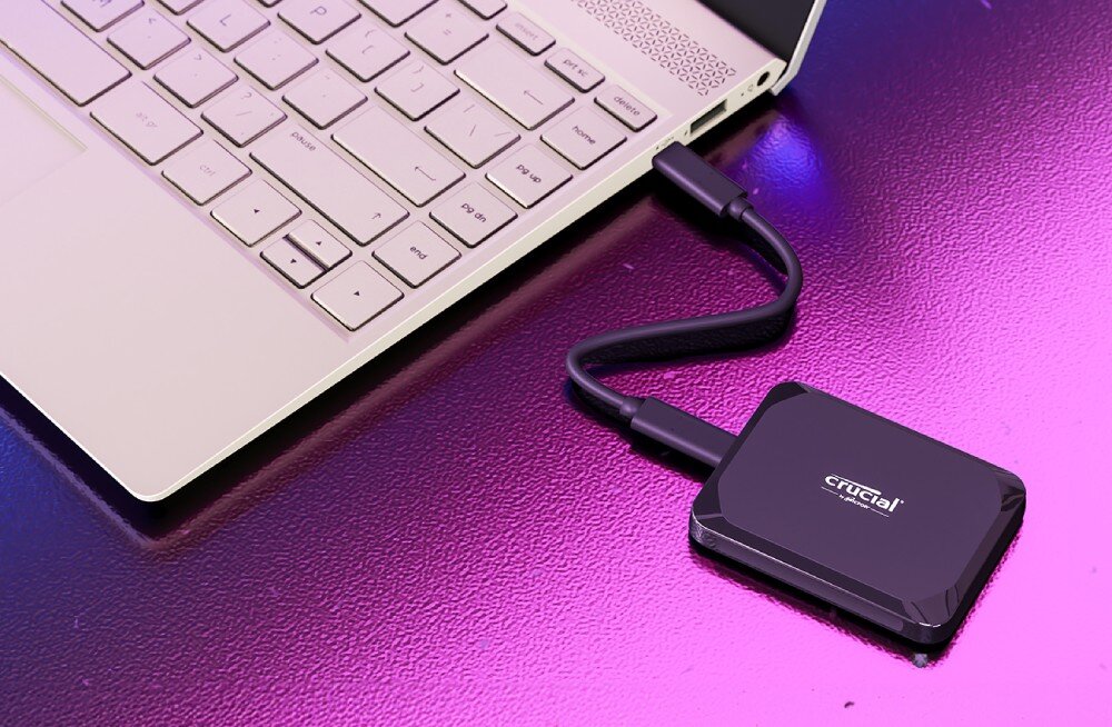 Dysk CRUCIAL X9 Przenośny dysk w czarnej obudowie podłączony kablem USB do jasnego komputera na błyszczącej fioletowej powierzchni o ziarnistej fakturze, prędkość, interfejs, pojemność
