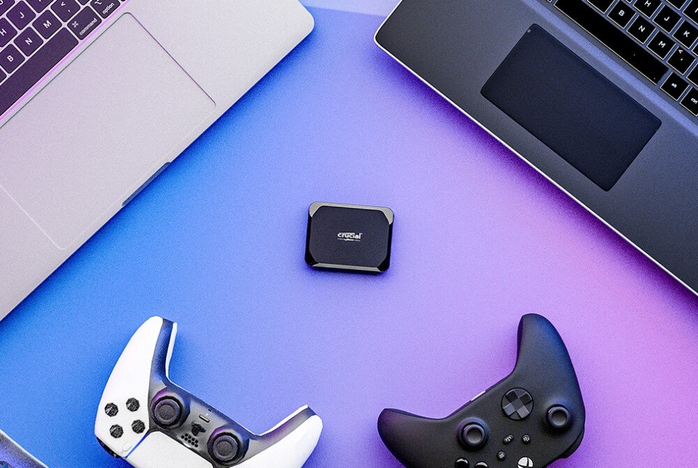 Dysk CRUCIAL X9 Niewielki czarny nośnik danych leżący centralnie na niebieskim tle pomiędzy dwoma kontrolerami do gier i narożnikami laptopów, kompatybilność, podłączenie, port