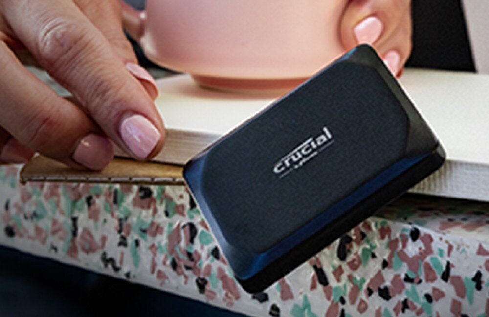 Dysk CRUCIAL X9 Kobieca dłoń z pomalowanymi paznokciami trzymająca czarne prostokątne urządzenie przy krawędzi jasnego przedmiotu, konstrukcja, odporność