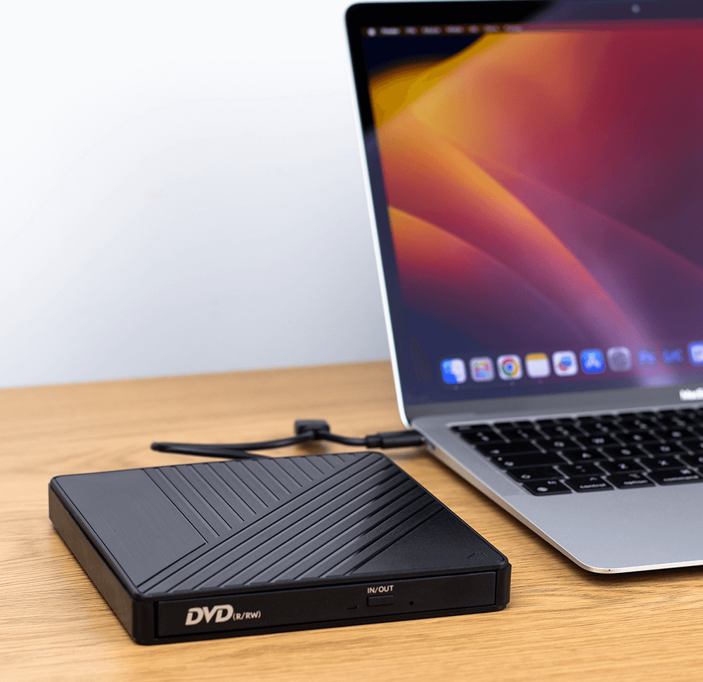 Na drewnianym blacie stoi czarny zewnętrzny napęd DVD z oznaczeniem \'DVD (R/RW)\' oraz laptop z otwartym ekranem. Napęd jest podłączony do laptopa za pomocą kabla USB.
