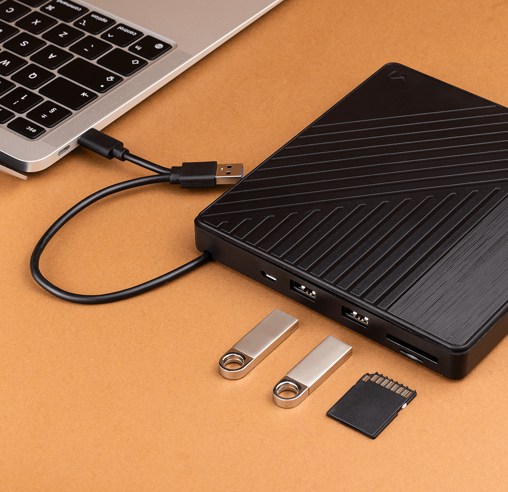 Na obrazie widoczny jest czarny zewnętrzny napęd z kilkoma portami USB, podłączony do laptopa za pomocą kabla USB. Obok napędu leżą dwa pendrive\'y i karta SD.