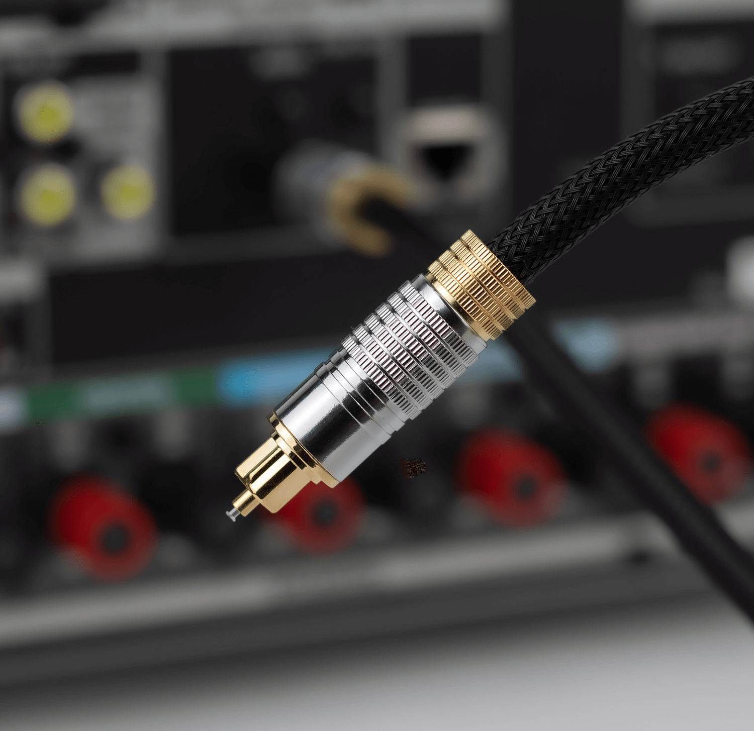 Zbliżenie na kabel optyczny z metalową końcówką w kolorze srebrnym i złotym, podłączony do urządzenia audio z widocznymi złączami i pokrętłami.