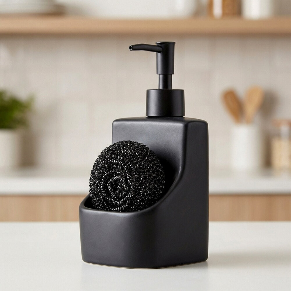 Czarny ceramiczny dozownik płynu do naczyń z pojemnikiem na gąbkę, w którym znajduje się czarna gąbka. Produkt stoi na jasnym blacie, a w tle widać rozmytą kuchnię.