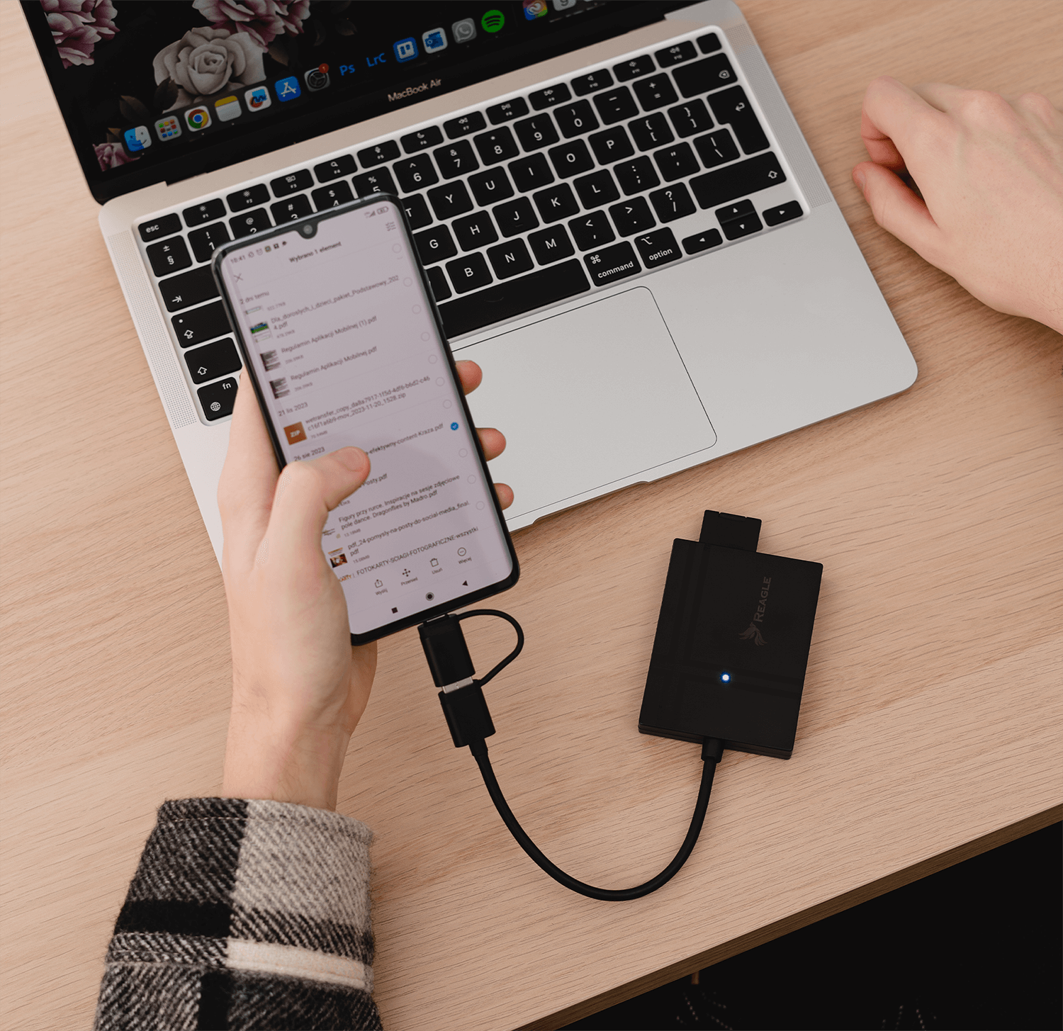 Osoba trzyma smartfon podłączony do czytnika kart pamięci, który leży obok laptopa na drewnianym biurku. Na czytniku widoczne jest logo \'REAGLE\'.
