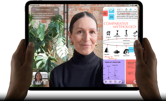 Widoczny od przodu ustawiony poziomo iPad Pro w kolorze gwiezdnej czerni, zaokrąglone rogi, czarna ramka wyświetlacza. Ręce trzymają urządzenie z lewej i prawej strony, na ekranie tłumaczona na żywo rozmowa przez FaceTime. Widać też aplikację edukacyjną z prezentacją „Comparative Mythology”, z tekstem, diagramami i odręcznymi notatkami