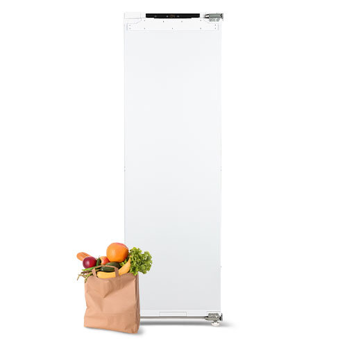 Lodówka, obok papierowa torba wypełniona warzywami i owocami.