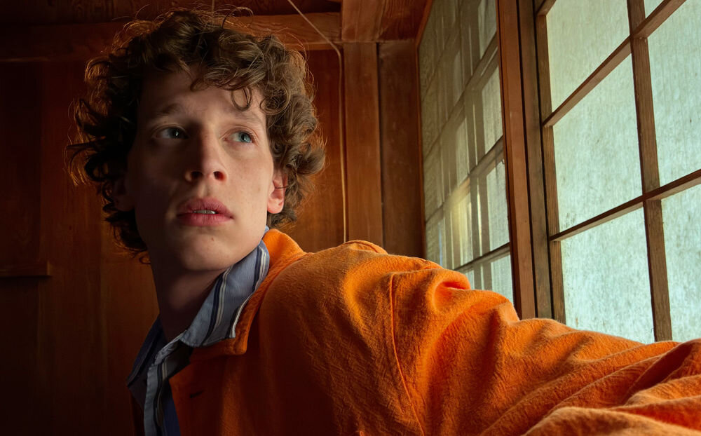 Smartfon APPLE iPhone 17e Portret młodego mężczyzny w pomarańczowej kurtce wykonany iPhone'em w trudnych warunkach oświetleniowych, prezentujący szczegółowość detali i naturalne odwzorowanie kolorów skóry przy świetle wpadającym przez okno.