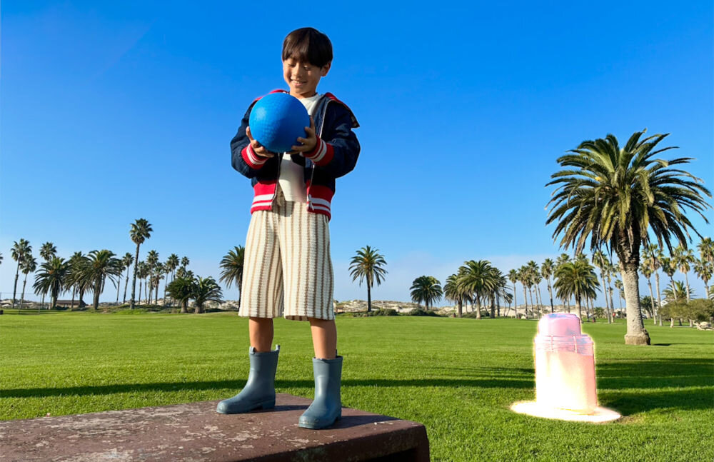 Smartfon APPLE iPhone 17e Zdjęcie dziecka trzymającego niebieską piłkę na trawiastym terenie z palmami. Po prawej stronie widoczny efekt inteligentnego wycinania obiektu z tła (funkcja Visual Look Up), podkreślający plecak z poświatą.