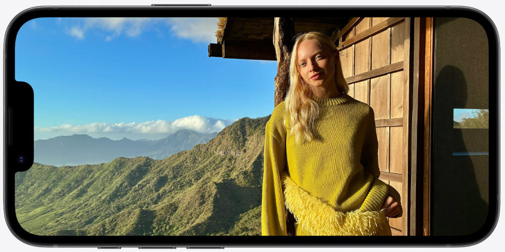 Smartfon APPLE iPhone 17e iPhone trzymany poziomo, wyświetlający wysokiej jakości zdjęcie portretowe kobiety w żółtym swetrze na tle malowniczego, górzystego krajobrazu pod błękitnym niebem.
