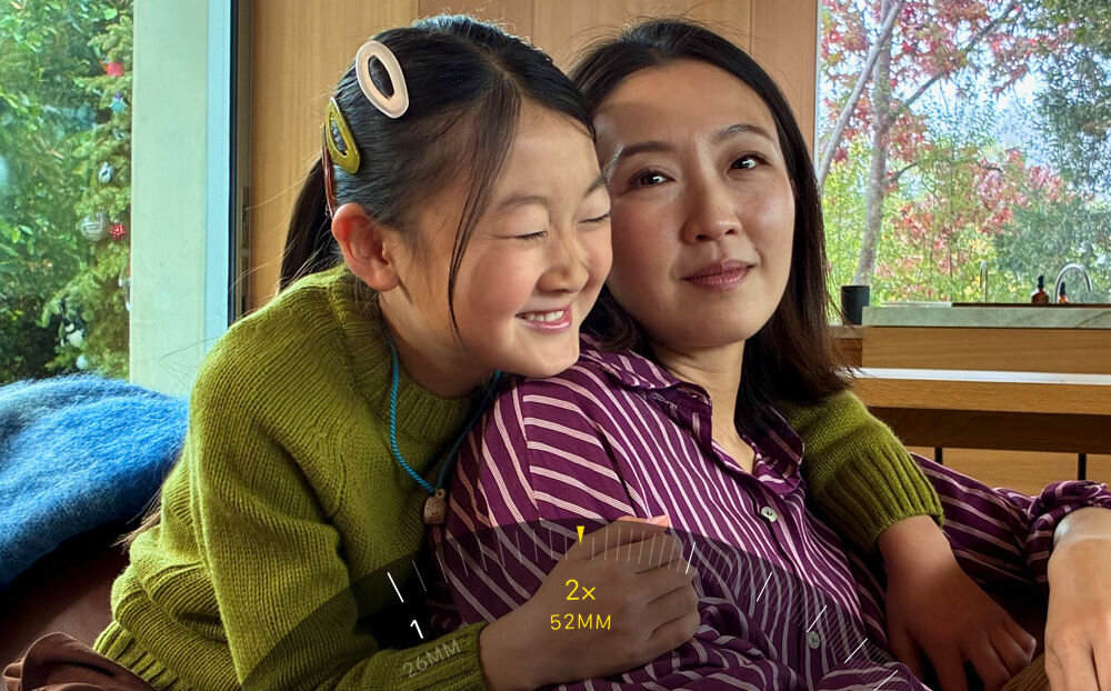 Smartfon APPLE iPhone 17e Zbliżenie na ekran iPhone'a podczas robienia zdjęcia. Interfejs aparatu pokazuje funkcję dwukrotnego zoomu (2x, 52mm) ustawioną na uśmiechniętą kobietę i dziewczynkę w domowym otoczeniu.