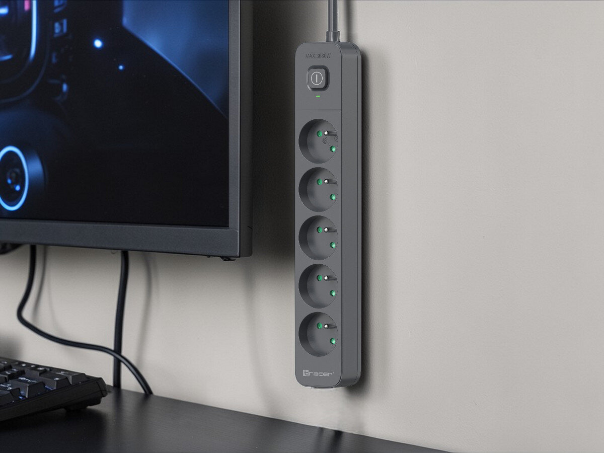 Na ścianie wisi czarna listwa zasilająca z sześcioma gniazdami, z zielonymi wskaźnikami LED. Obok znajduje się monitor i klawiatura, a na listwie widoczne jest logo \'Tracer\'.