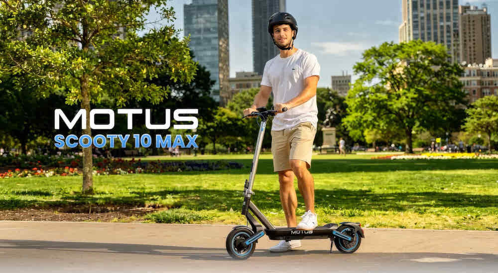 Hulajnoga elektryczna MOTUS Scooty 10 Max 80km 800W Amortyzacja 10 cali park osoba opiera się o hulajnogę w tle wysokie budynki napis MOTUS Scooty 10 Max środek transportu styl stabilność