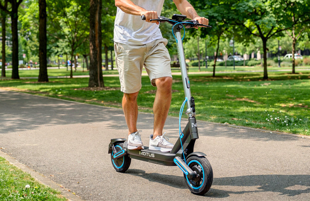 Hulajnoga elektryczna MOTUS Scooty 10 Max 80km 800W Amortyzacja 10 cali park osoba jedzie na hulajnodze po drodze w tle ludzie drzewa koła bezdętkowe o średnicy 10 cali niski opór toczenia duża przyczepność do podłoża płynna i oszczędna jazda absorbują wstrząsy i nierówności terenu opony są odporne na przebicia i nie wymagają konserwacji