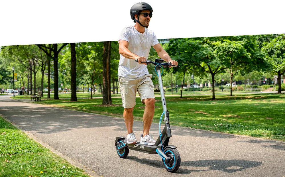 Hulajnoga elektryczna MOTUS Scooty 10 Max 80km 800W Amortyzacja 10 cali miasto osoba jedzie na hulajnodze po chodniku w tle ludzie drzewa system KERS odzyskiwanie energii podczas hamowania i jazdy z górki energia elektryczna wykorzystywana ponownie w trakcie jazdy zwiększa się efektywność hulajnogi system działa automatycznie