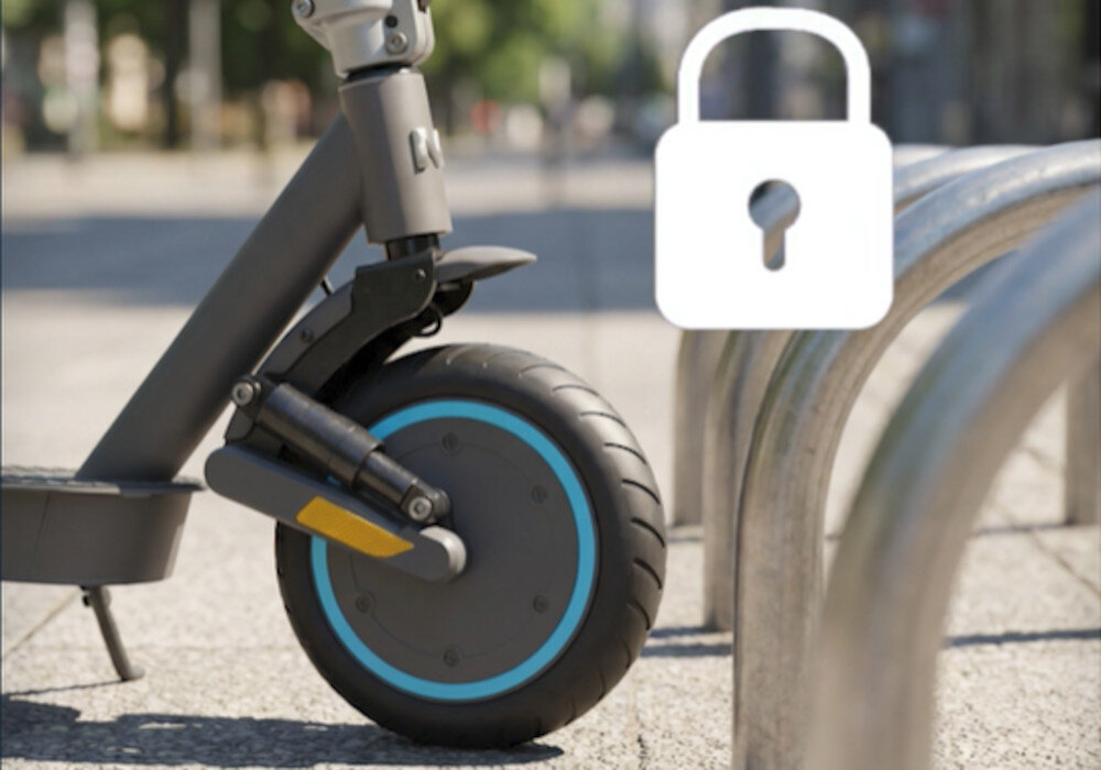 Zbliżenie na przednie koło hulajnogi elektrycznej z amortyzacją, obok którego znajduje się symbol kłódki. W tle widoczne są metalowe barierki na chodniku.