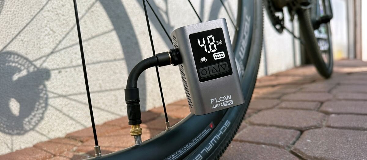 Pompka rowerowa podłączona do wentyla koła rowerowego, z wyświetlaczem pokazującym 4.8 BAR oraz ikoną roweru. Na urządzeniu widoczny jest napis \'FLOW AIR12 PRO\'.