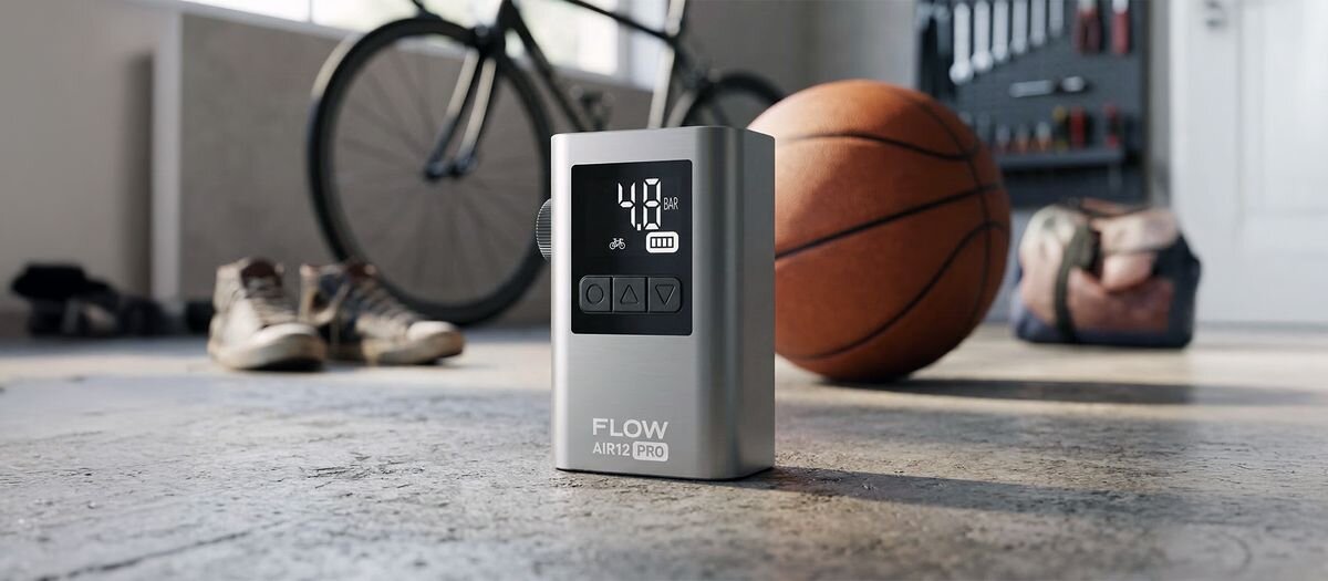 Na podłodze stoi pompka rowerowa z wyświetlaczem pokazującym \'40 psi\' oraz ikoną baterii. W tle widoczne są rower, piłka do koszykówki i para butów.