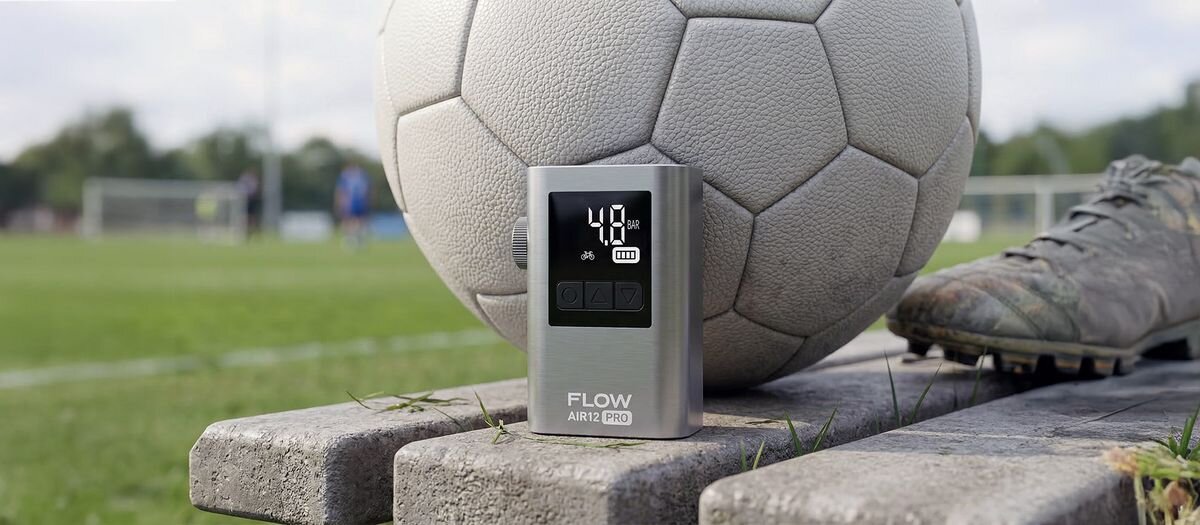 Na drewnianej ławce znajduje się pompka rowerowa z wyświetlaczem pokazującym \'4.0\' oraz piłka nożna. Na pompce widoczny jest napis \'FLOW AIR12 PRO\'.