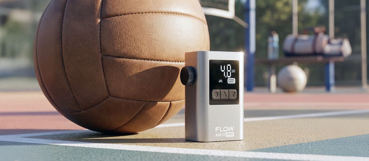 Na boisku sportowym znajduje się pompka rowerowa z wyświetlaczem pokazującym \'40 PSI\' oraz piłka do siatkówki. Na pompce widoczny jest napis \'FLOW AIR12 PRO\'.